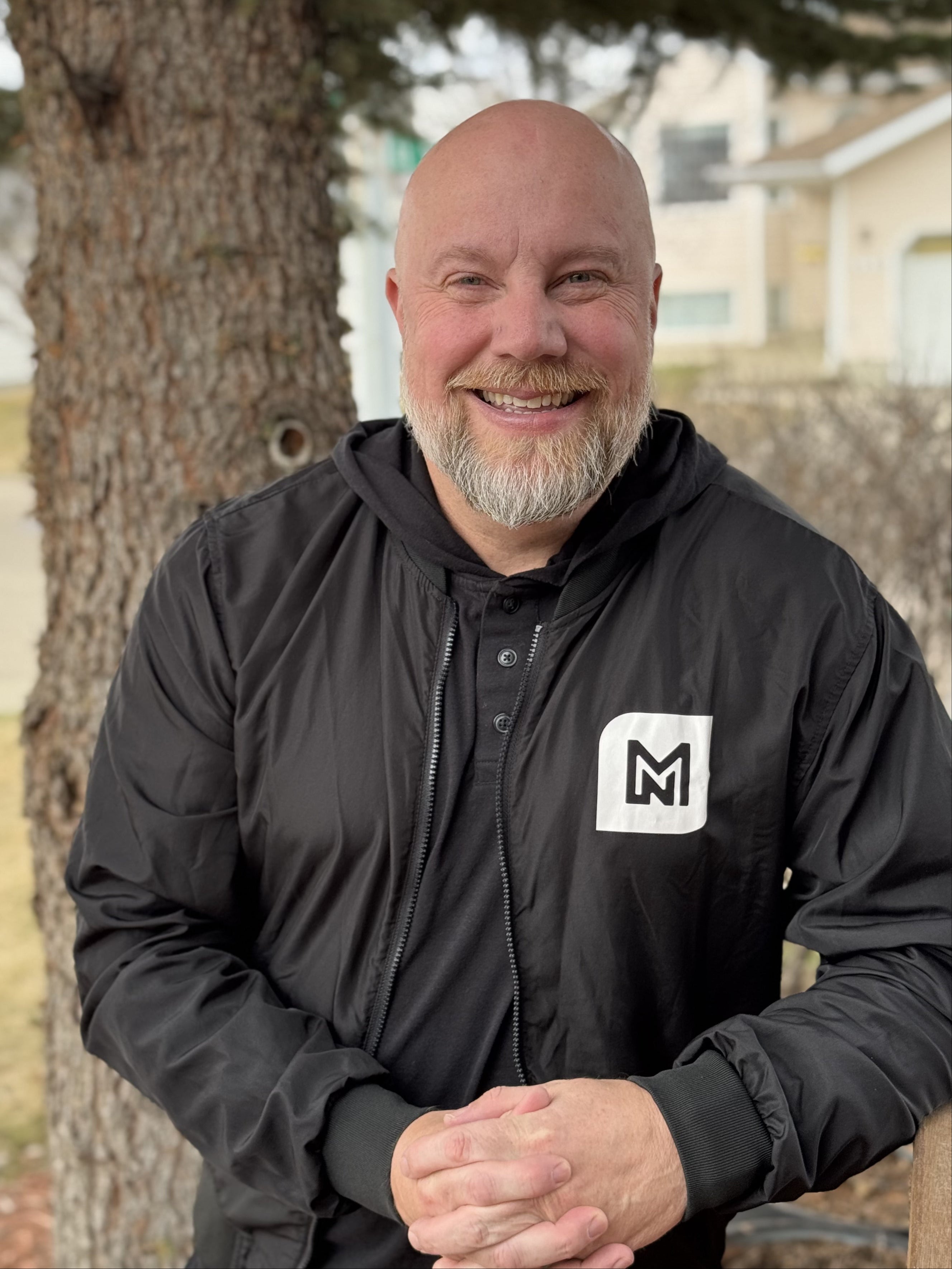 Paul Fraser - Man wearing a black jacket with a logo standing outdoors next to a tree